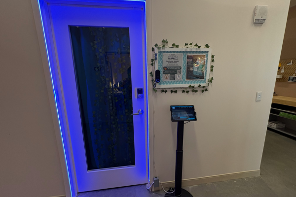 Exterior office door framed by electric blue RGBW LED strip with ivy curtain visible through glass, Reolink PoE doorbell camera mounted to branded bulletin board, and Galaxy tablet on floor stand kiosk