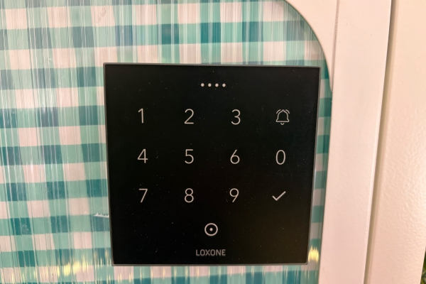 Close-up of Loxone NFC Touch Air keypad mounted on cabinet door against teal gingham background, showing numeric touch interface with bell and checkmark icons and Loxone branding