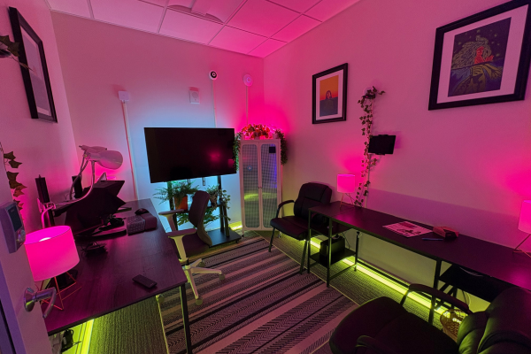 Full showroom interior in party mode with dynamic pink and magenta Hue lighting saturating the ceiling and walls, green RGBW floor perimeter LED strips, glowing cabinet with plants, TV rack, two desks, and three framed artwork pieces